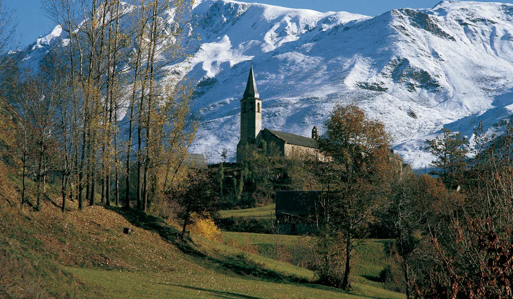 Vista exterior de l'església de Santa Eulària d'Unha amb arbres al davant i la muntanya nevada al darrere. 